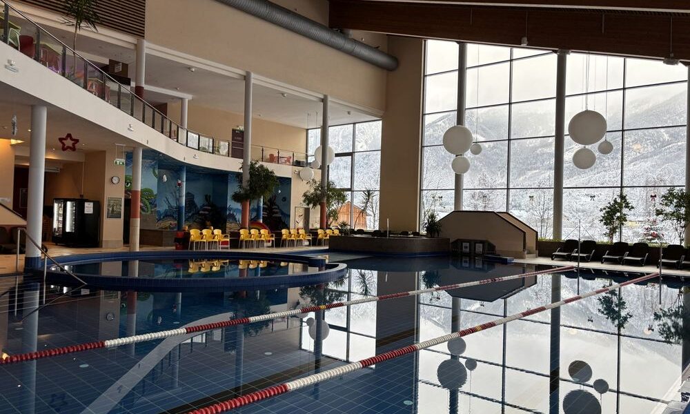 Hallenbad mit Bahntrennung, große Fenster mit Blick auf die verschneite Landschaft draußen und Sitzbereiche mit gelben Stühlen an einer Seite.