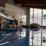 Hallenbad mit Bahntrennung, große Fenster mit Blick auf die verschneite Landschaft draußen und Sitzbereiche mit gelben Stühlen an einer Seite.