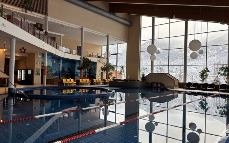 Hallenbad mit Bahntrennung, große Fenster mit Blick auf die verschneite Landschaft draußen und Sitzbereiche mit gelben Stühlen an einer Seite.