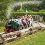 Ein kleiner Zug im Freien, der Erwachsene und Kinder befördert, überquert eine Holzbrücke inmitten von Grün auf einer Miniatureisenbahnstrecke.