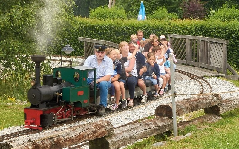 Ein kleiner Zug im Freien, der Erwachsene und Kinder befördert, überquert eine Holzbrücke inmitten von Grün auf einer Miniatureisenbahnstrecke.
