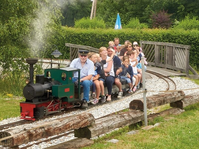 Ein kleiner Zug im Freien, der Erwachsene und Kinder befördert, überquert eine Holzbrücke inmitten von Grün auf einer Miniatureisenbahnstrecke.