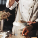 Ein Koch in weißer Uniform streut Gewürze über eine Metallschüssel mit Lebensmitteln, während eine andere Person den Inhalt in einer Küchenumgebung mischt.