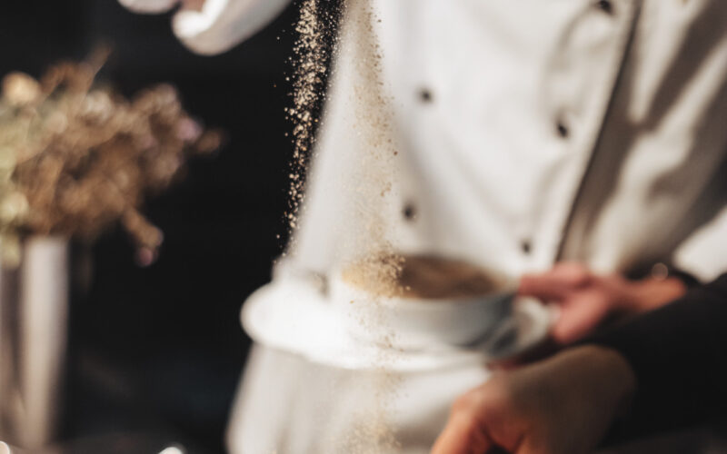 Ein Koch in weißer Uniform streut Gewürze über eine Metallschüssel mit Lebensmitteln, während eine andere Person den Inhalt in einer Küchenumgebung mischt.