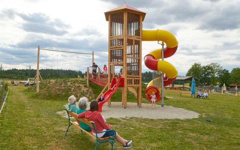 Kinder spielen auf einem hölzernen Spielplatz mit Rutschen, während drei Erwachsene auf einer Bank in der Nähe in einem grasbewachsenen Park unter einem bewölkten Himmel sitzen und zusehen.