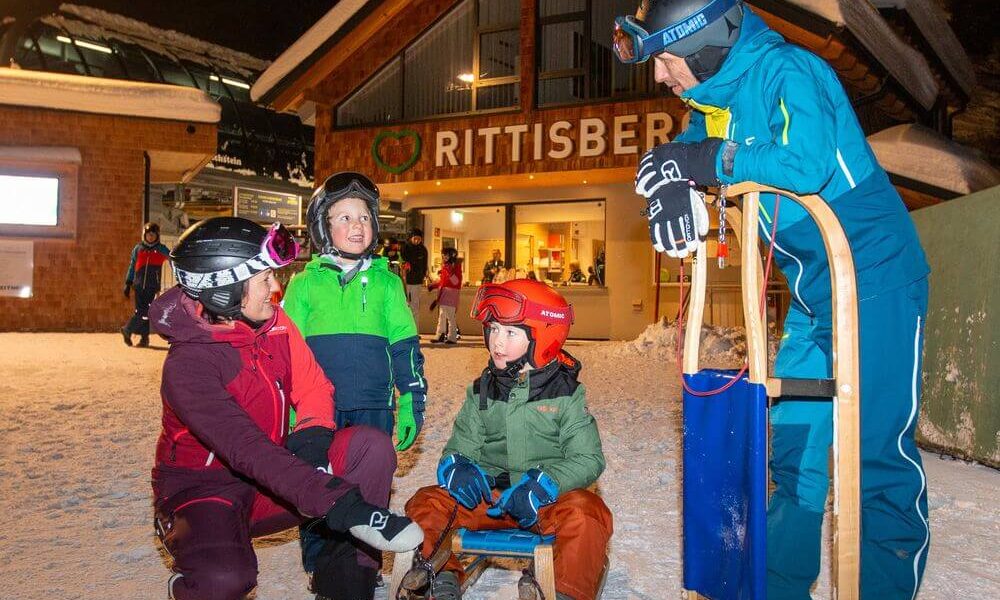 Vier Personen in Winterkleidung versammeln sich vor einem Gebäude mit der Aufschrift "Rittisberg"; zwei Kinder sitzen auf einem Schlitten, zwei Erwachsene stehen und knien in der Nähe.