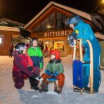 Vier Personen in Winterkleidung versammeln sich vor einem Gebäude mit der Aufschrift "Rittisberg"; zwei Kinder sitzen auf einem Schlitten, zwei Erwachsene stehen und knien in der Nähe.