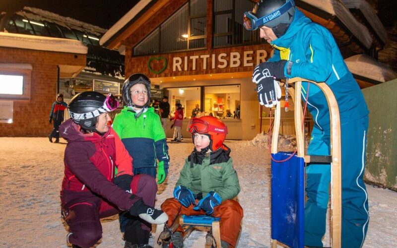 Vier Personen in Winterkleidung versammeln sich vor einem Gebäude mit der Aufschrift "Rittisberg"; zwei Kinder sitzen auf einem Schlitten, zwei Erwachsene stehen und knien in der Nähe.