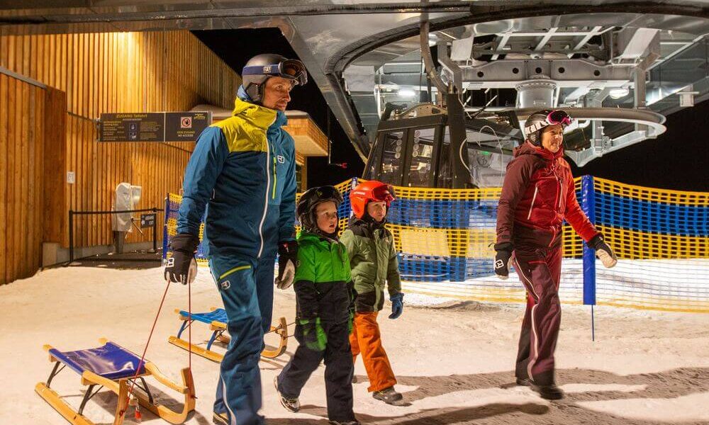 Eine vierköpfige Familie in Winterkleidung und mit Helmen geht nachts mit Schlitten in der Nähe eines Skilifts in einem verschneiten Skigebiet spazieren.