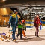 Eine vierköpfige Familie in Winterkleidung und mit Helmen geht nachts mit Schlitten in der Nähe eines Skilifts in einem verschneiten Skigebiet spazieren.