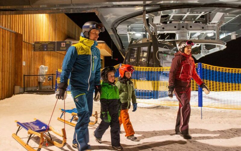 Eine vierköpfige Familie in Winterkleidung und mit Helmen geht nachts mit Schlitten in der Nähe eines Skilifts in einem verschneiten Skigebiet spazieren.