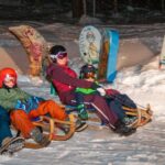 Vier Personen, zwei Erwachsene und zwei Kinder, rodeln nachts einen verschneiten Hügel hinunter, mit Helmen und Winterkleidung, mit bunten Brettern im Hintergrund.