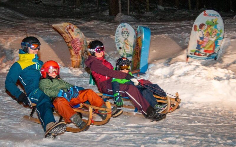 Vier Personen, zwei Erwachsene und zwei Kinder, rodeln nachts einen verschneiten Hügel hinunter, mit Helmen und Winterkleidung, mit bunten Brettern im Hintergrund.