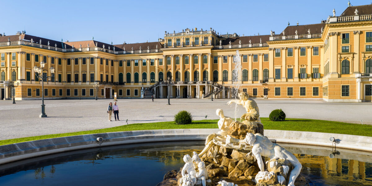 Barockpalast mit gelber Fassade, großen Fenstern und verzierten Statuen, die einen Brunnen im Vordergrund unter blauem Himmel umgeben.