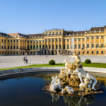 Barockpalast mit gelber Fassade, großen Fenstern und verzierten Statuen, die einen Brunnen im Vordergrund unter blauem Himmel umgeben.