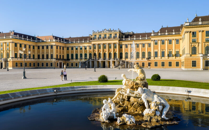 Barockpalast mit gelber Fassade, großen Fenstern und verzierten Statuen, die einen Brunnen im Vordergrund unter blauem Himmel umgeben.
