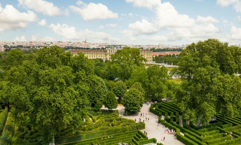 Ein formaler Garten mit geometrischen Hecken und Wegen, großen grünen Bäumen und Spaziergängern, mit einem historischen Gebäude und einer Stadtlandschaft im Hintergrund unter einem teilweise bewölkten Himmel.