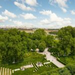 Ein formaler Garten mit geometrischen Hecken und Wegen, großen grünen Bäumen und Spaziergängern, mit einem historischen Gebäude und einer Stadtlandschaft im Hintergrund unter einem teilweise bewölkten Himmel.