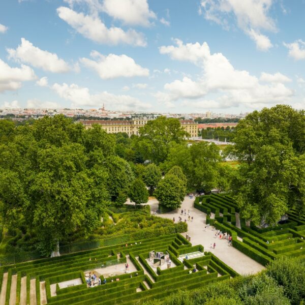 Ein formaler Garten mit geometrischen Hecken und Wegen, großen grünen Bäumen und Spaziergängern, mit einem historischen Gebäude und einer Stadtlandschaft im Hintergrund unter einem teilweise bewölkten Himmel.