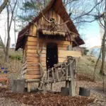 Ein kleines hölzernes Spielhaus mit einem steilen Dach und einem rustikalen Holzzaun steht zwischen blattlosen Bäumen auf einem gemulchten Boden.