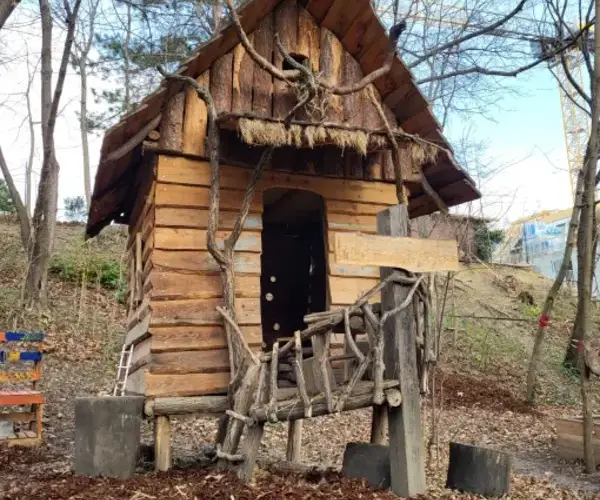 Ein kleines hölzernes Spielhaus mit einem steilen Dach und einem rustikalen Holzzaun steht zwischen blattlosen Bäumen auf einem gemulchten Boden.
