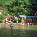 Kinder spielen in einem flachen Wasserbereich in der Nähe eines bunten Zugwagens; Erwachsene und andere Kinder sind in der Nähe unter Bäumen versammelt.