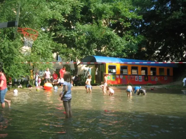 Kinder spielen in einem flachen Wasserbereich in der Nähe eines bunten Zugwagens; Erwachsene und andere Kinder sind in der Nähe unter Bäumen versammelt.