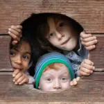 Drei Kinder spähen durch ein rundes Loch in einer Holzwand und schauen mit neugierigen und verspielten Gesichtern hinaus.