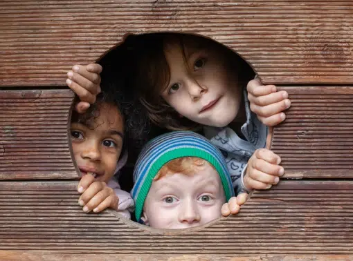 Drei Kinder spähen durch ein rundes Loch in einer Holzwand und schauen mit neugierigen und verspielten Gesichtern hinaus.