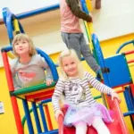 Drei kleine Kinder spielen auf bunten Indoor-Spielgeräten; ein Mädchen rutscht eine rote Rutsche hinunter, während zwei andere auf der Struktur klettern.