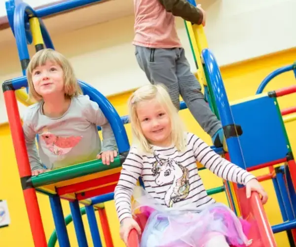 Drei kleine Kinder spielen auf bunten Indoor-Spielgeräten; ein Mädchen rutscht eine rote Rutsche hinunter, während zwei andere auf der Struktur klettern.