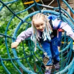 Ein Kind mit geflochtenem Haar krabbelt durch einen blauen Seiltunnel in einem von Bäumen umgebenen Outdoor-Abenteuerpark.