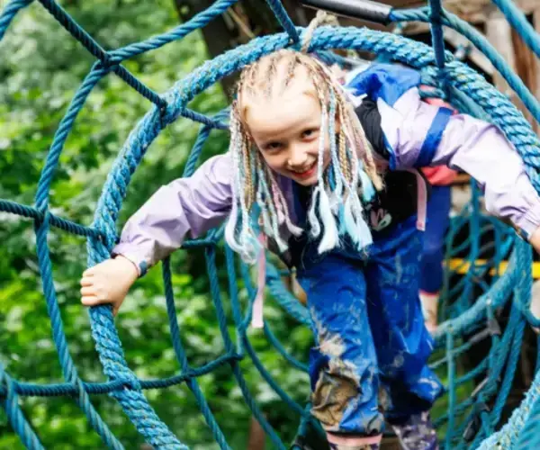 Ein Kind mit geflochtenem Haar krabbelt durch einen blauen Seiltunnel in einem von Bäumen umgebenen Outdoor-Abenteuerpark.