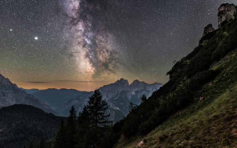 Die Milchstraße ist an einem klaren Nachthimmel über Berggipfeln und einem bewaldeten Hügel zu sehen.
