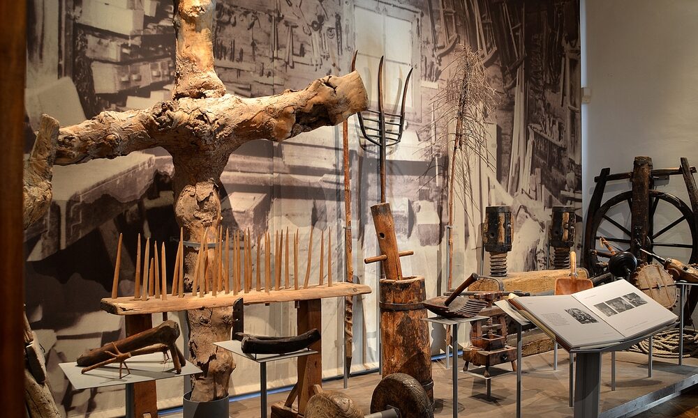 In einer Museumsausstellung sind alte landwirtschaftliche Werkzeuge, Geräte und Maschinenteile aus Holz vor dem Hintergrund eines großen historischen Schwarz-Weiß-Fotos zu sehen.
