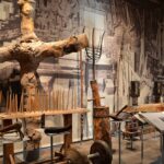 In einer Museumsausstellung sind alte landwirtschaftliche Werkzeuge, Geräte und Maschinenteile aus Holz vor dem Hintergrund eines großen historischen Schwarz-Weiß-Fotos zu sehen.