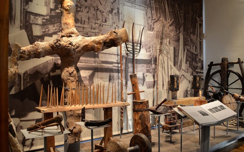 In einer Museumsausstellung sind alte landwirtschaftliche Werkzeuge, Geräte und Maschinenteile aus Holz vor dem Hintergrund eines großen historischen Schwarz-Weiß-Fotos zu sehen.