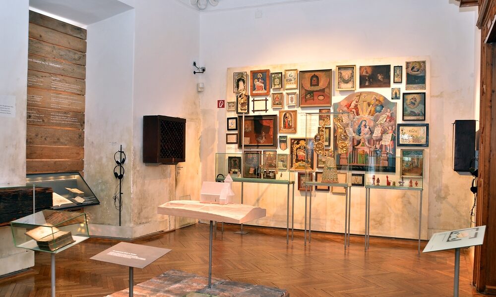 In einem Ausstellungsraum des Museums sind gerahmte Gemälde an der Wand, Glasvitrinen mit Artefakten und Dokumenten sowie Holzböden zu sehen.