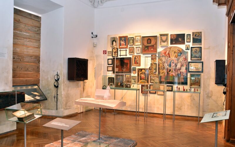 In einem Ausstellungsraum des Museums sind gerahmte Gemälde an der Wand, Glasvitrinen mit Artefakten und Dokumenten sowie Holzböden zu sehen.