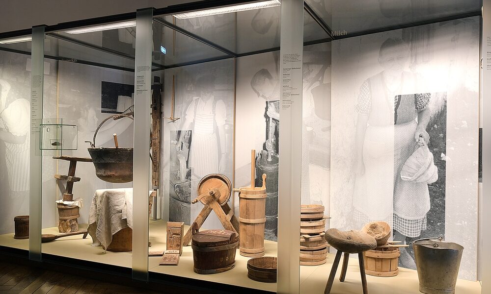 Eine Museumsvitrine zeigt alte hölzerne Molkereigeräte und -ausrüstungen, darunter Kannen, Eimer, Hocker und Schaufeln, mit Schwarz-Weiß-Fotos von Menschen, die sie benutzen, im Hintergrund.