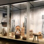 Eine Museumsvitrine zeigt alte hölzerne Molkereigeräte und -ausrüstungen, darunter Kannen, Eimer, Hocker und Schaufeln, mit Schwarz-Weiß-Fotos von Menschen, die sie benutzen, im Hintergrund.