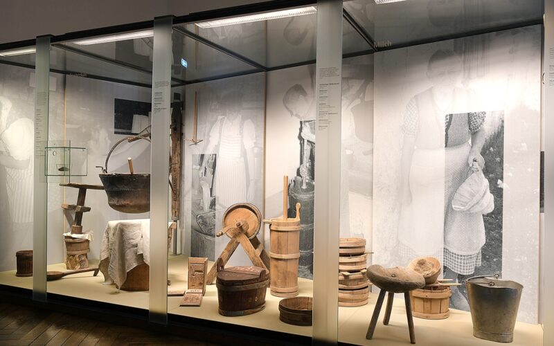 Eine Museumsvitrine zeigt alte hölzerne Molkereigeräte und -ausrüstungen, darunter Kannen, Eimer, Hocker und Schaufeln, mit Schwarz-Weiß-Fotos von Menschen, die sie benutzen, im Hintergrund.