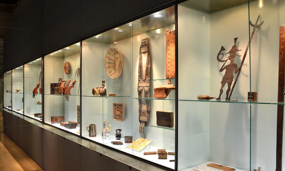 Eine Museumsvitrine mit verschiedenen historischen Artefakten, darunter Töpferwaren, Metallarbeiten, geschnitzte Gegenstände und Figuren, die auf Glasregalen mit Oberlicht angeordnet sind.