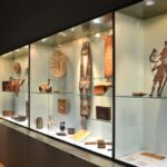 Eine Museumsvitrine mit verschiedenen historischen Artefakten, darunter Töpferwaren, Metallarbeiten, geschnitzte Gegenstände und Figuren, die auf Glasregalen mit Oberlicht angeordnet sind.