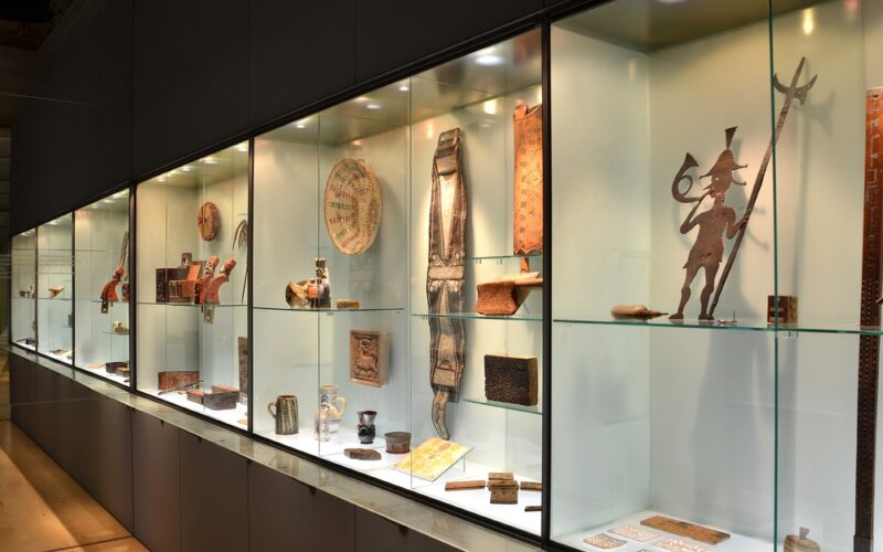 Eine Museumsvitrine mit verschiedenen historischen Artefakten, darunter Töpferwaren, Metallarbeiten, geschnitzte Gegenstände und Figuren, die auf Glasregalen mit Oberlicht angeordnet sind.