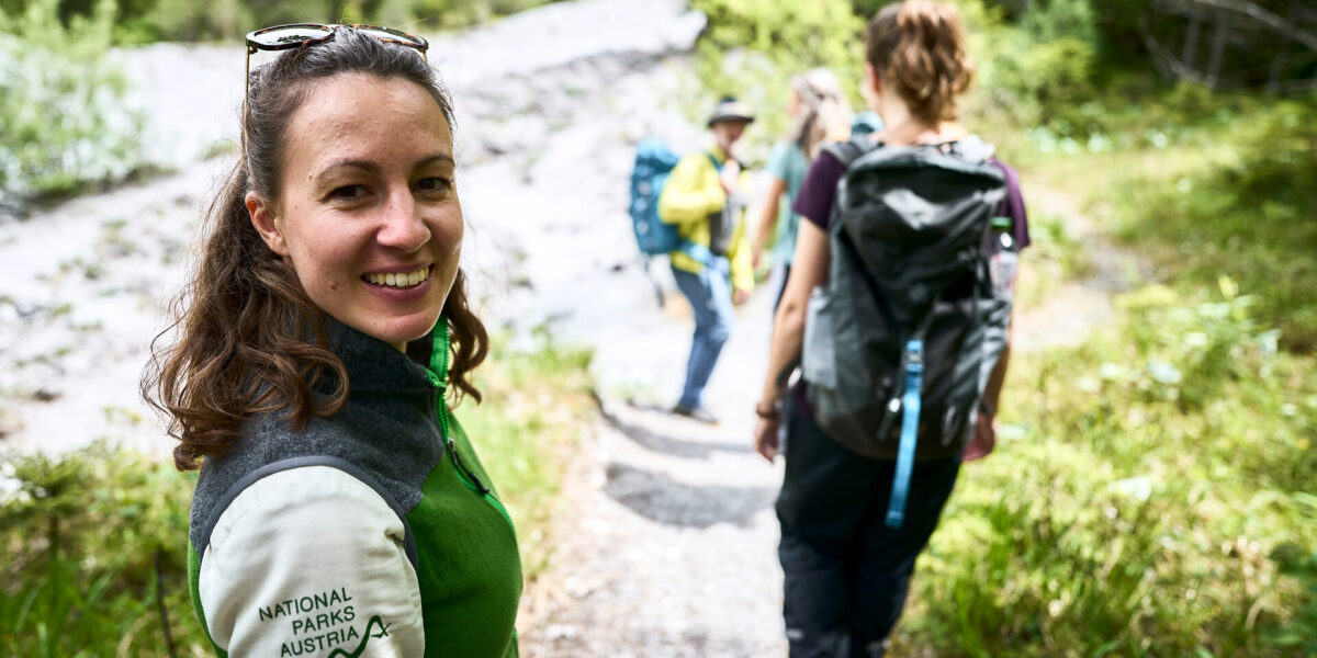 Eine Frau mit einer "Nationalparks Austria"-Jacke lächelt in die Kamera, während sie mit drei anderen auf einem Waldweg wandert.