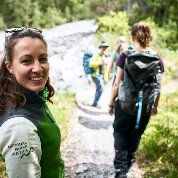 Eine Frau mit einer "Nationalparks Austria"-Jacke lächelt in die Kamera, während sie mit drei anderen auf einem Waldweg wandert.
