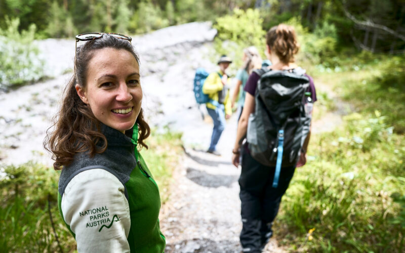 Eine Frau mit einer "Nationalparks Austria"-Jacke lächelt in die Kamera, während sie mit drei anderen auf einem Waldweg wandert.