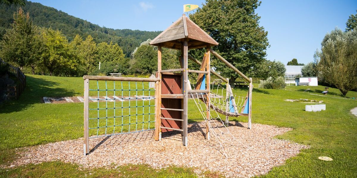 Ein hölzernes Spielgerät mit Kletternetzen und einer kleinen Rutsche steht auf einer Wiese mit Bäumen und Hügeln im Hintergrund.