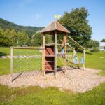 Ein hölzernes Spielgerät mit Kletternetzen und einer kleinen Rutsche steht auf einer Wiese mit Bäumen und Hügeln im Hintergrund.